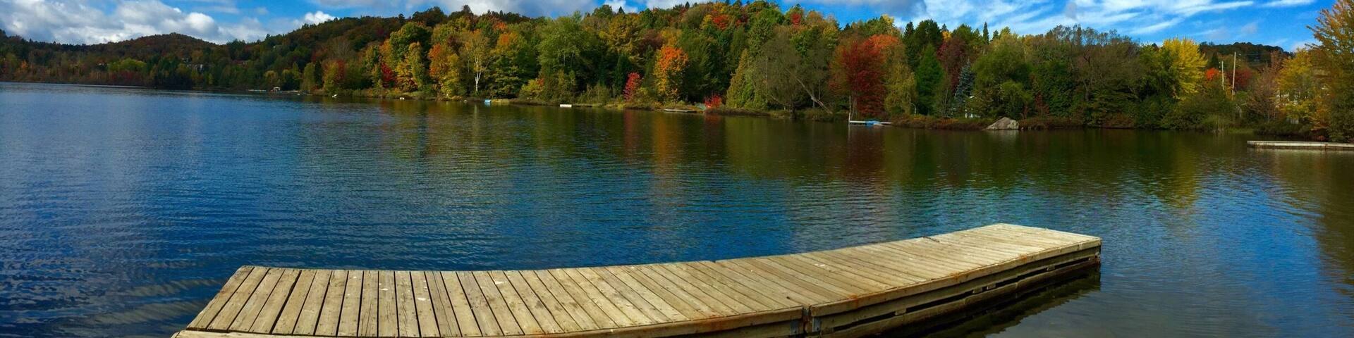 A wooden dock extending into a calm lake with autumn-colored trees in the background and a partly cloudy sky.