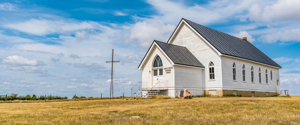 A white church with a cross in a grassy field under a blue sky.