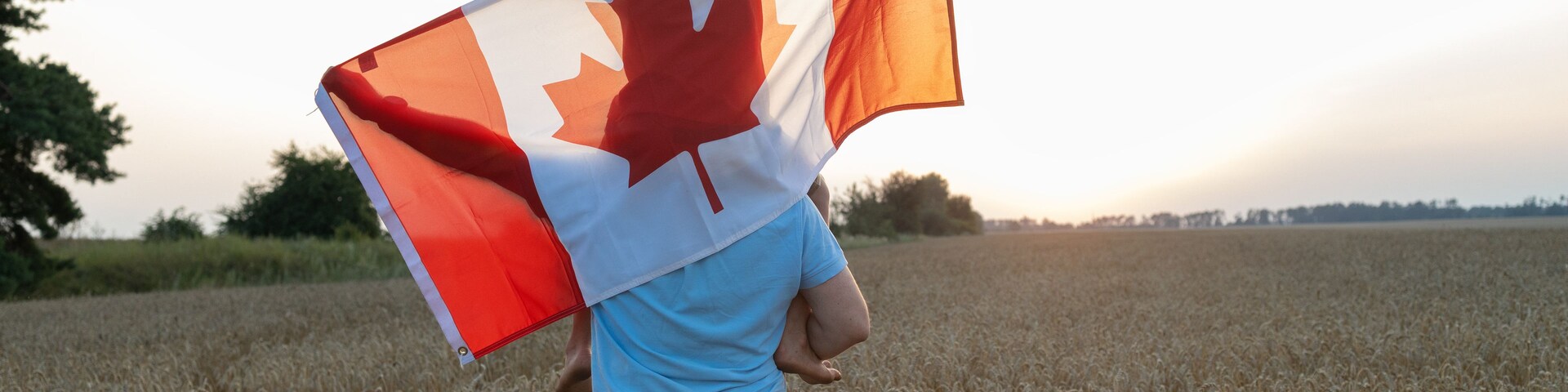 large Canadian flag draped over boy sitting on shoulders of man, standing in a wheat field at sunset. Happy Canada Day Pride, freedom, patriotism. A journey across the country. A national symbol