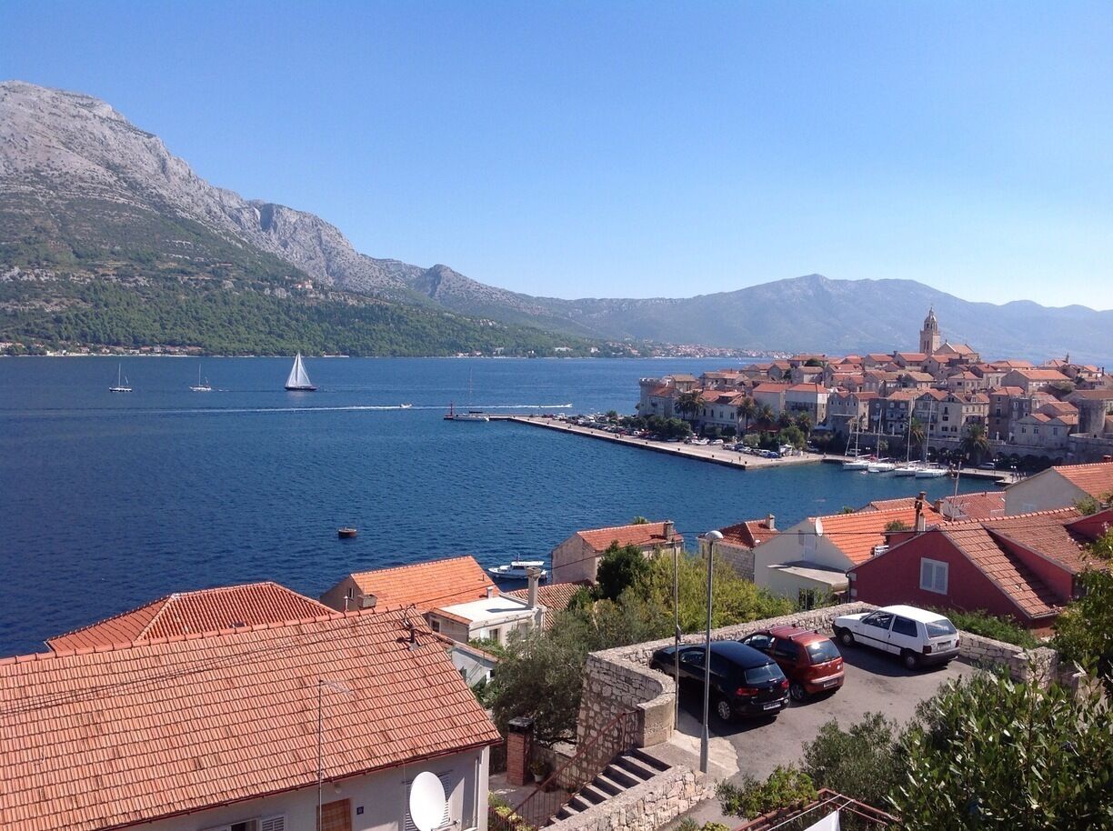A coastal town with red-roofed buildings, a marina with sailboats, and mountains in the background.