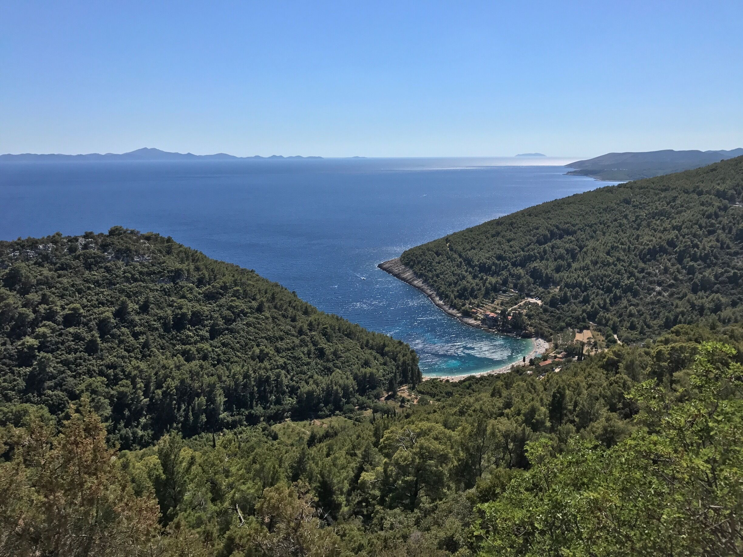 A coastal landscape with dense forests, a clear blue sea, and distant mountains.