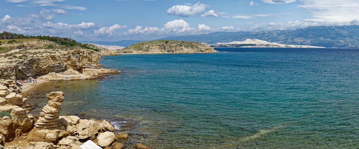 A coastal landscape with clear blue waters, rocky cliffs, and distant snow-capped mountains.