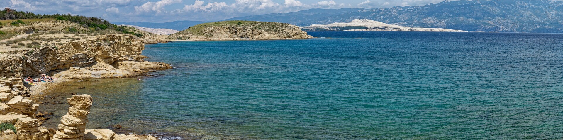 A coastal landscape with clear blue waters, rocky cliffs, and distant snow-capped mountains.