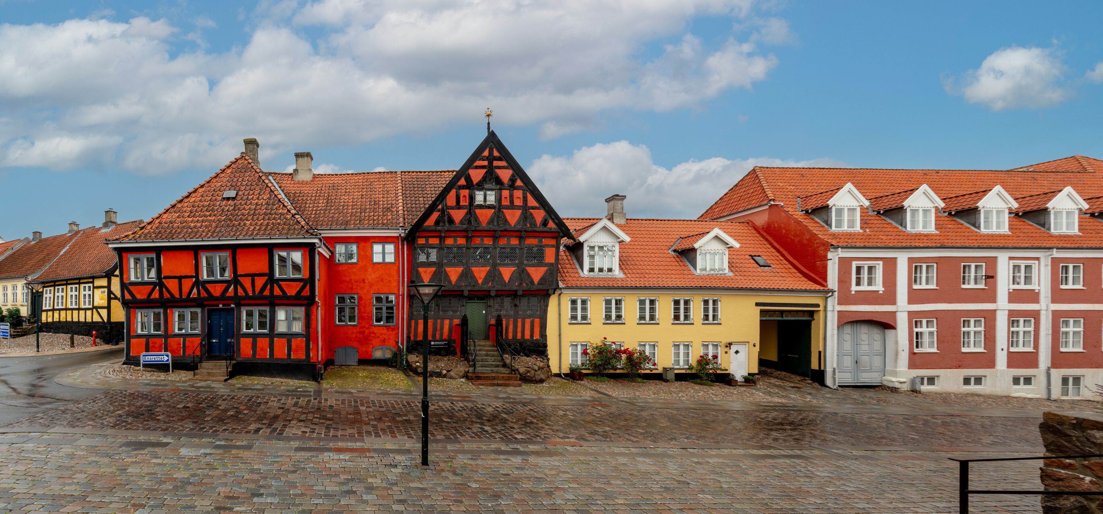 A row of colorful half-timbered houses with red and yellow facades, a cobblestone street, and a clear blue sky.
