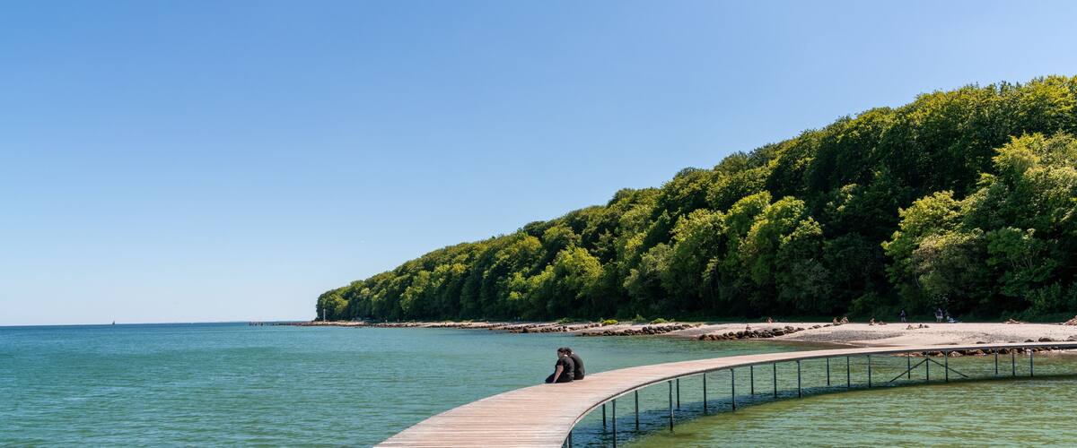 A wooden boardwalk curving along a beach with a forested hill in the background.