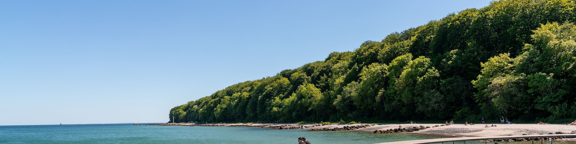 A wooden boardwalk curving along a beach with a forested hill in the background.
