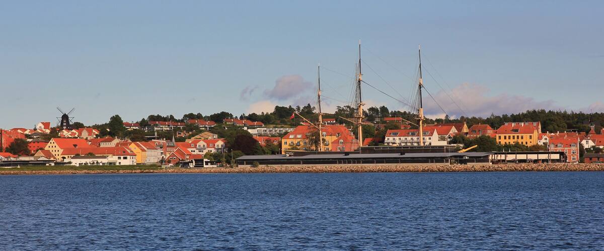 A coastal town with red-roofed houses, a windmill, and a large ship with tall masts.
