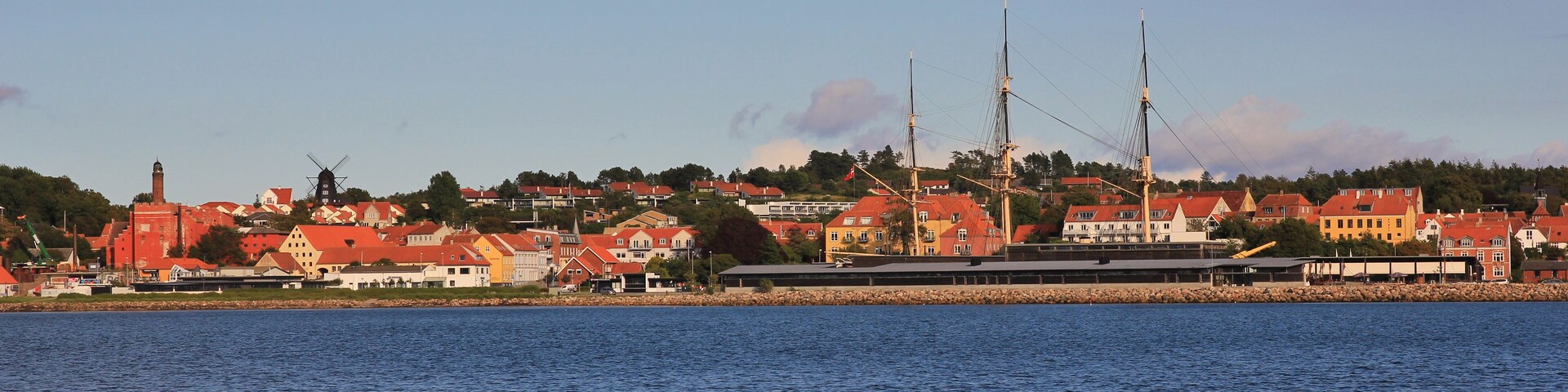 A coastal town with red-roofed houses, a windmill, and a large ship with tall masts.
