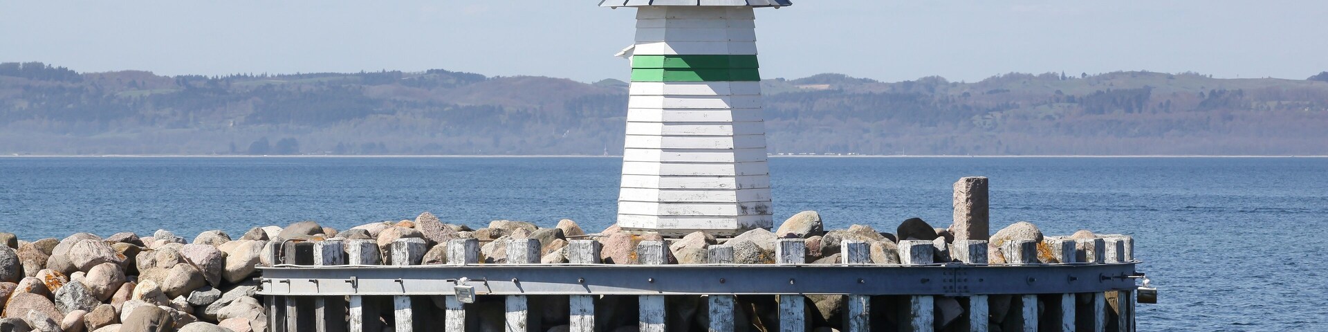 Um farol sobre um cais rochoso, com vista clara para a água e colinas ao longe.