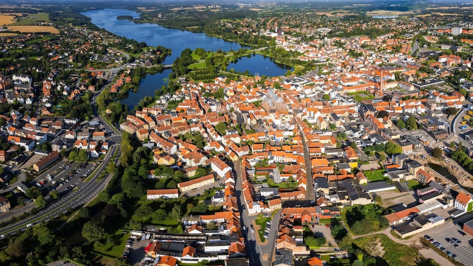 An aerial view of a town with a river, surrounded by green fields and red-roofed buildings.