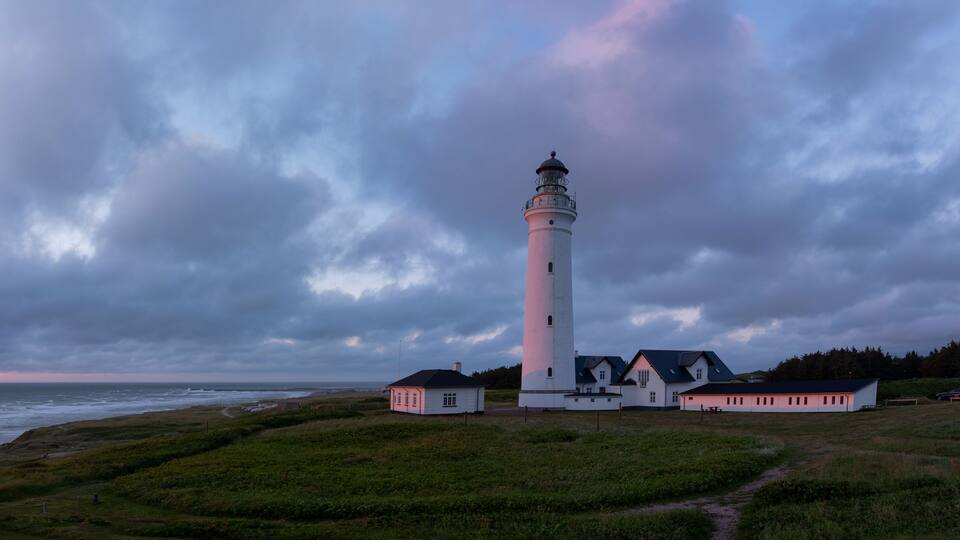 A white lighthouse with a black top, surrounded by a small white building with a red roof, set against a cloudy sky.