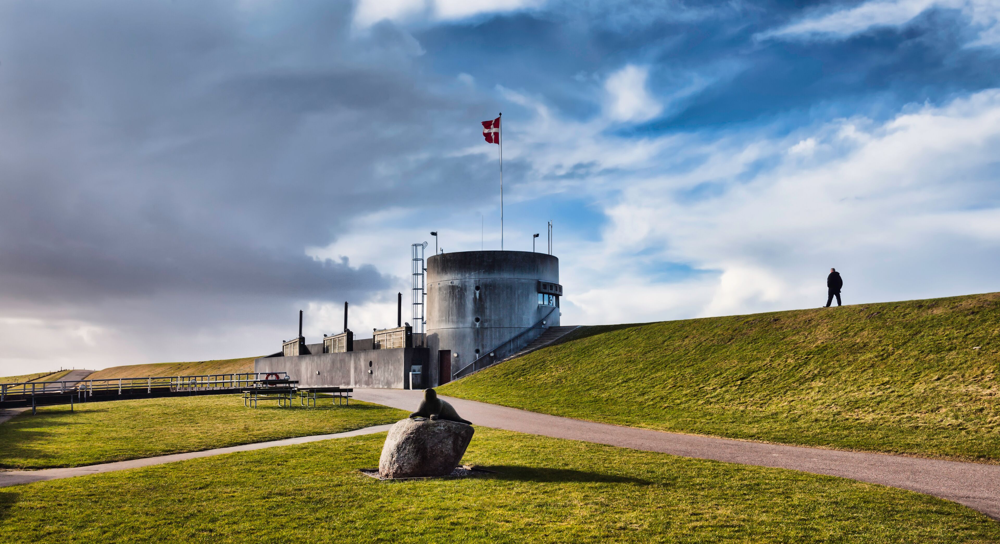 A grassy hill with a flagpole, a large cylindrical building, and a person walking.