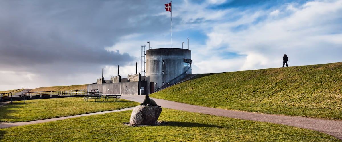 A grassy hill with a flagpole, a large cylindrical building, and a person walking.