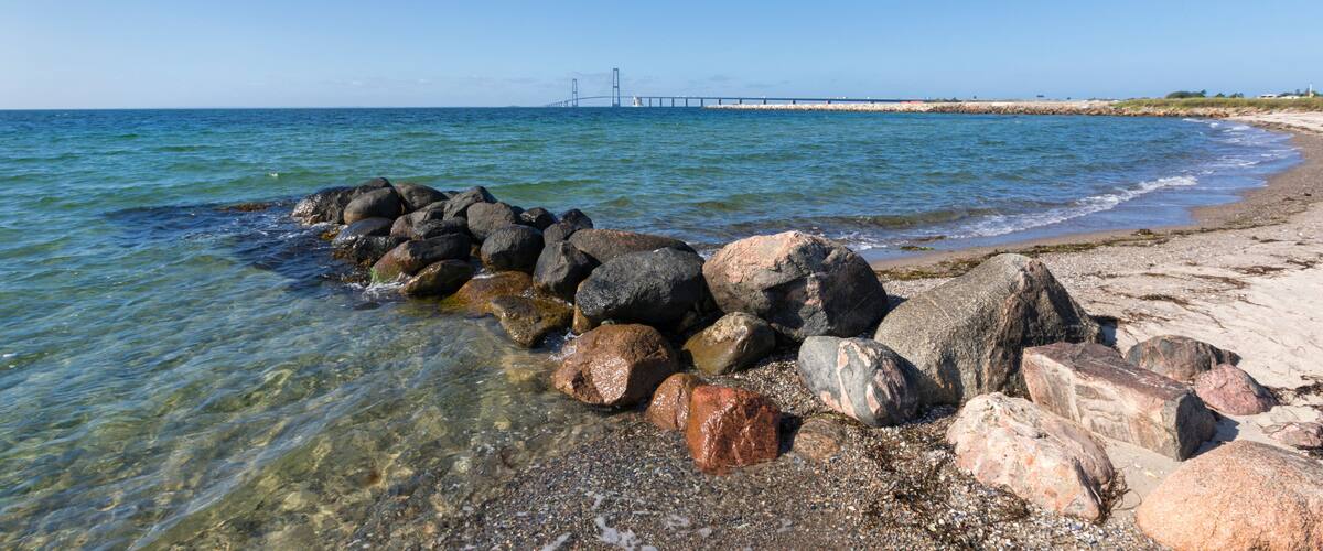 A rocky shoreline with clear water, a distant bridge, and a lighthouse.