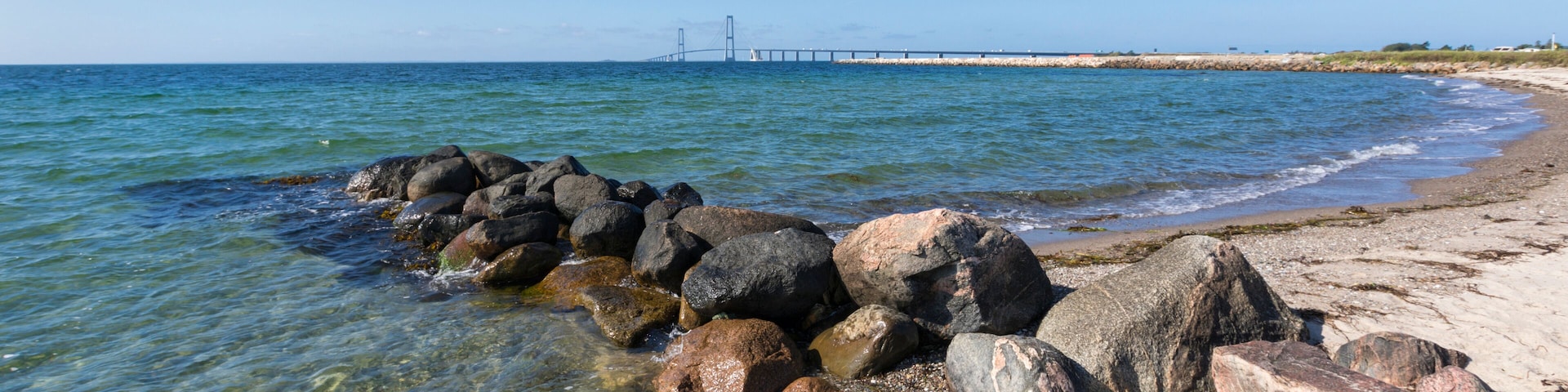A rocky shoreline with clear water, a distant bridge, and a lighthouse.