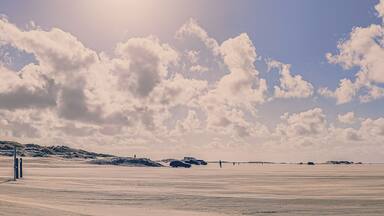 A vast desert landscape with rolling dunes, a clear sky with scattered clouds, and a few vehicles and people in the distance.