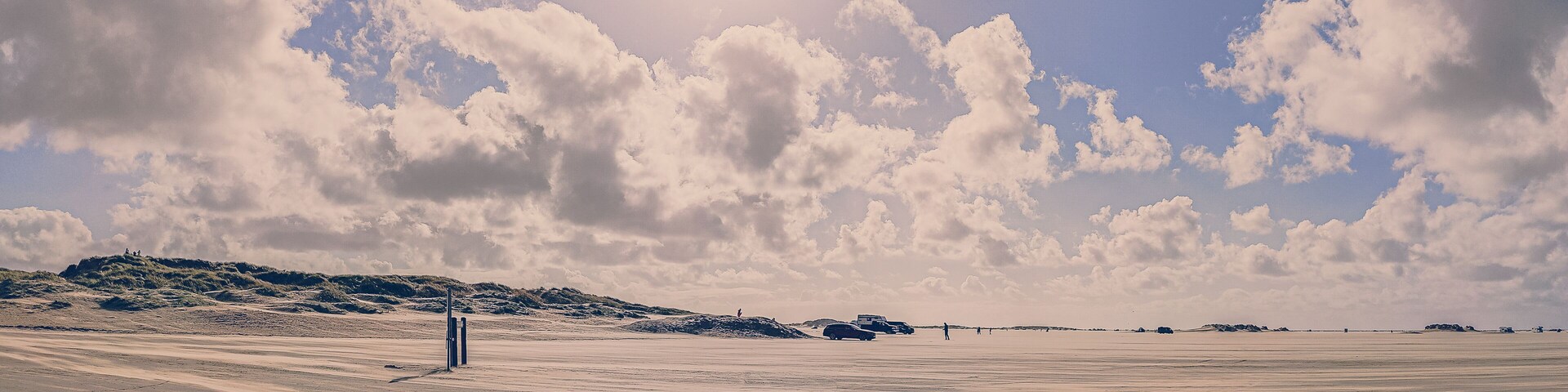 A vast desert landscape with rolling dunes, a clear sky with scattered clouds, and a few vehicles and people in the distance.