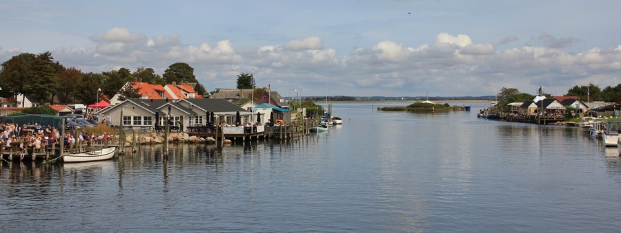 A calm waterfront with docked boats, a pier with people, and buildings with red roofs.