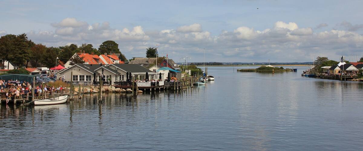 A calm waterfront with docked boats, a pier with people, and buildings with red roofs.