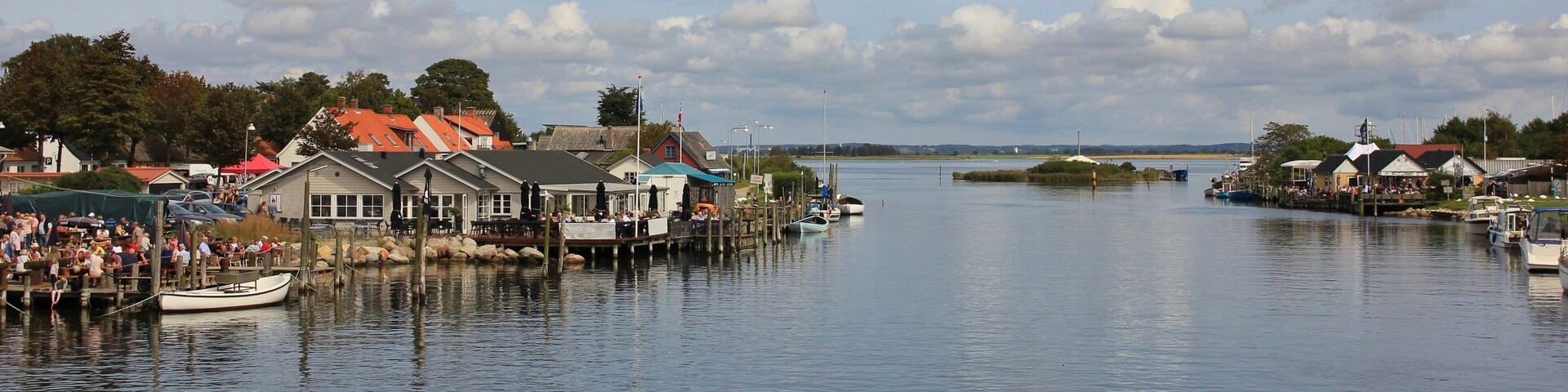 A calm waterfront with docked boats, a pier with people, and buildings with red roofs.