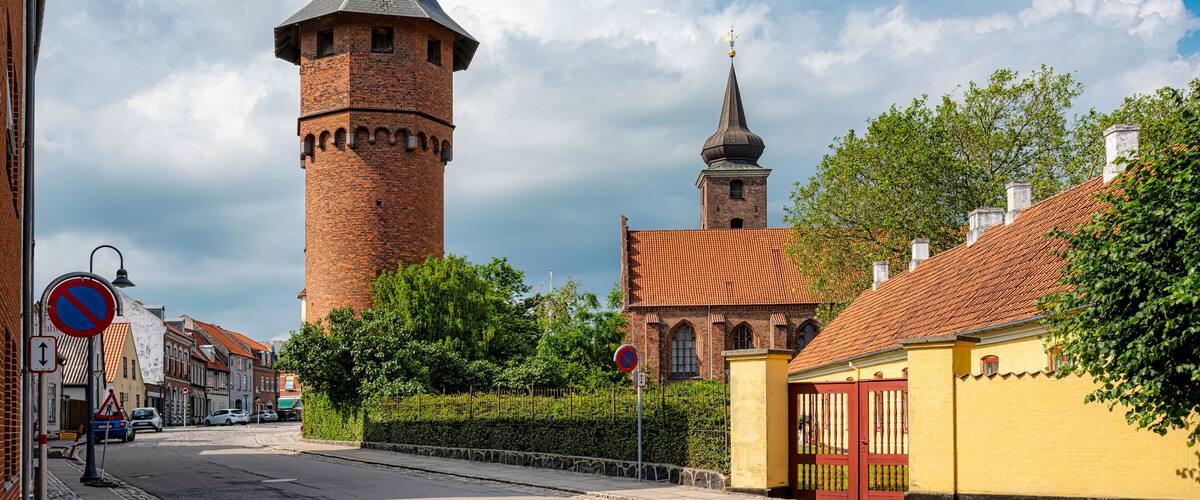 A historic tower with a pointed roof, a church with a dome, and a yellow building with a red gate.