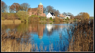 A serene lakeside village with a prominent church tower, traditional houses, and a calm water body reflecting the architecture.