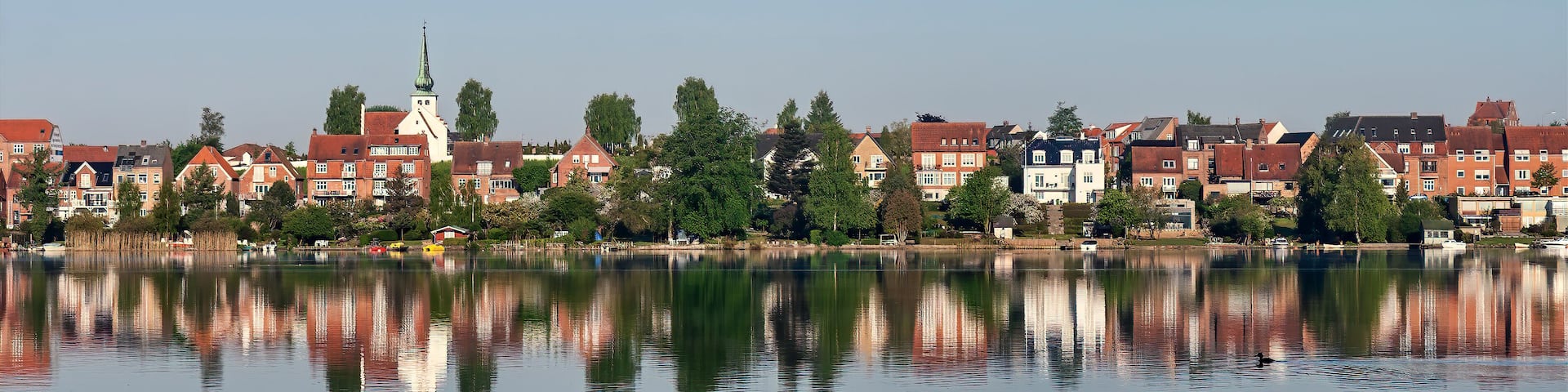 A calm lake reflecting buildings and a church steeple.