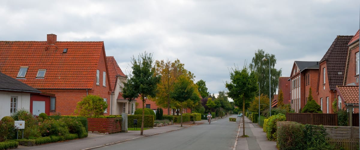 A residential street lined with red-tiled houses, a cyclist, and a cloudy sky.