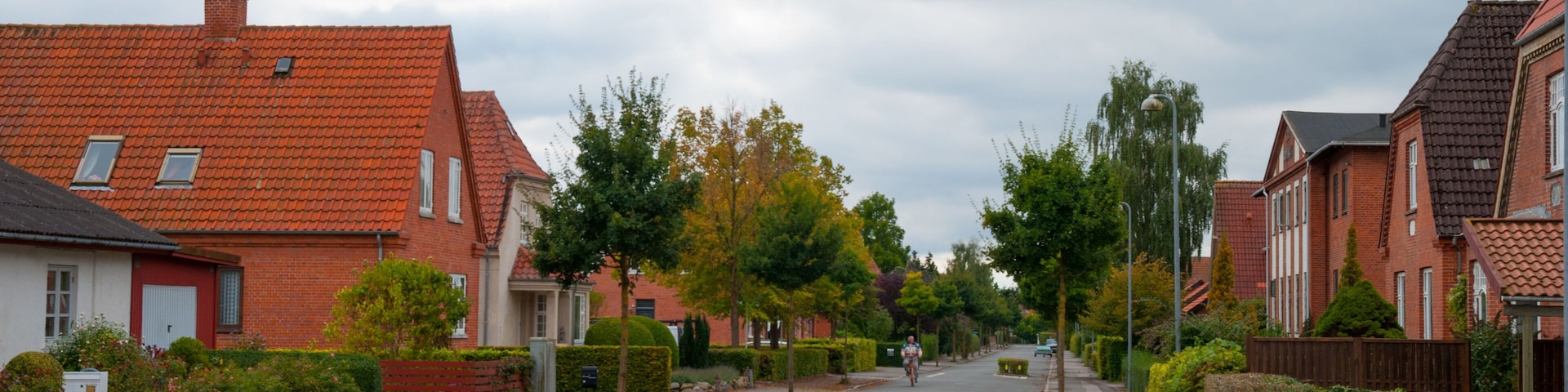 A residential street lined with red-tiled houses, a cyclist, and a cloudy sky.