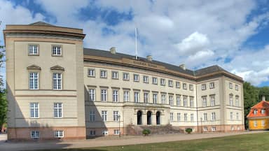 A large academic building with a red-roofed annex, surrounded by a well-maintained lawn and a lake.