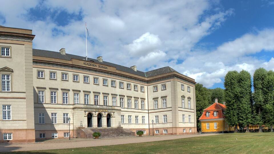 A large academic building with a red-roofed annex, surrounded by a well-maintained lawn and a lake.