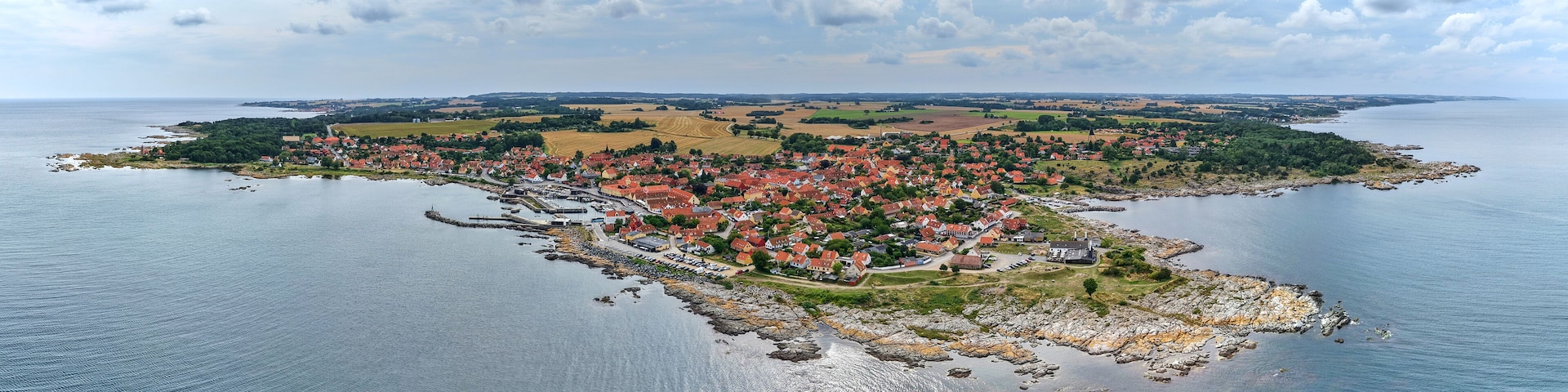 An aerial view of a coastal town with red-roofed houses, a harbor, and surrounding fields.