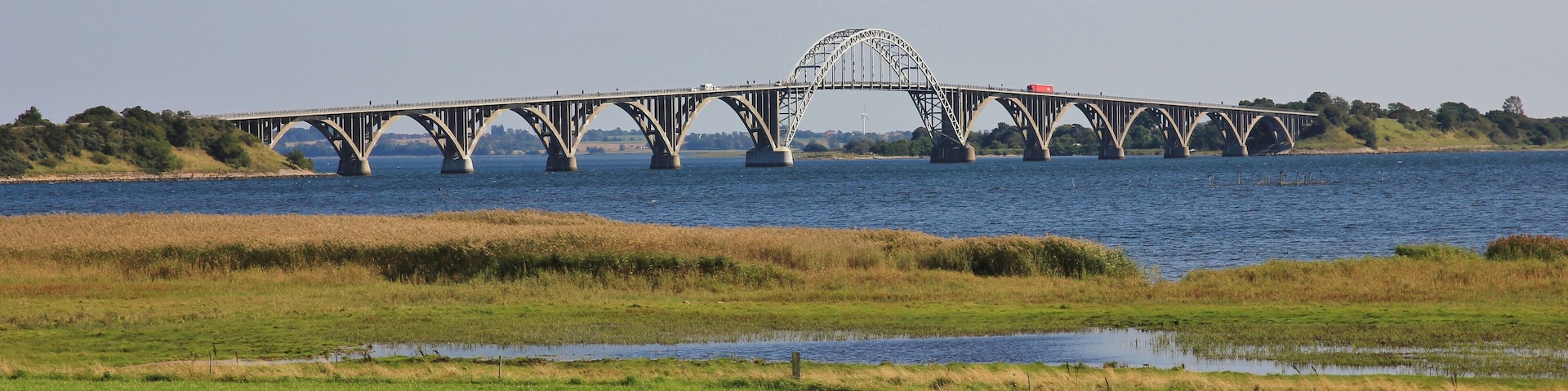 A large steel bridge spanning a river with grassy banks and a small body of water in the foreground.