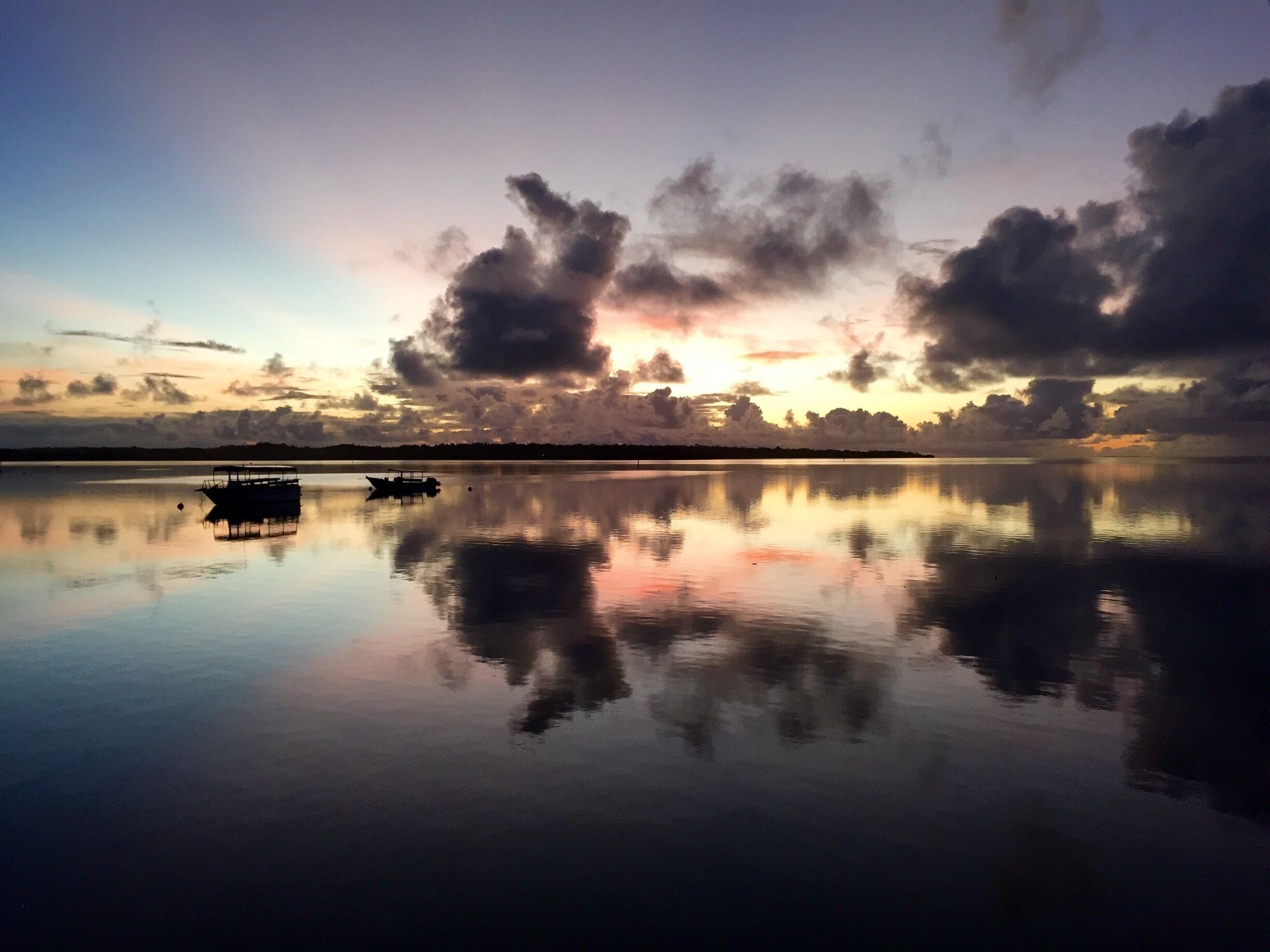 A calm lake at sunset with a boat in the foreground and a silhouette of trees on the horizon.