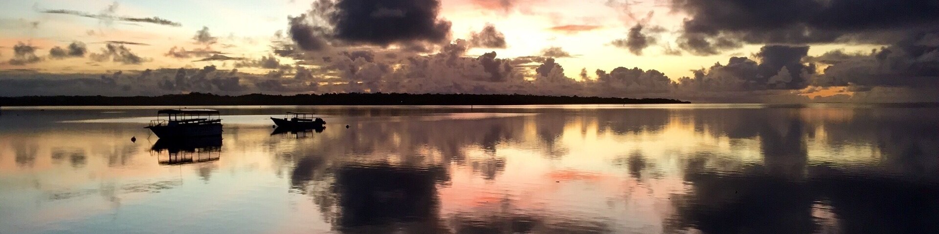 A calm lake at sunset with a boat in the foreground and a silhouette of trees on the horizon.