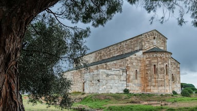 Ein historisches Steinhaus mit Rundbogenfenstern und flachem Dach, umgeben von Gras und Bäumen.