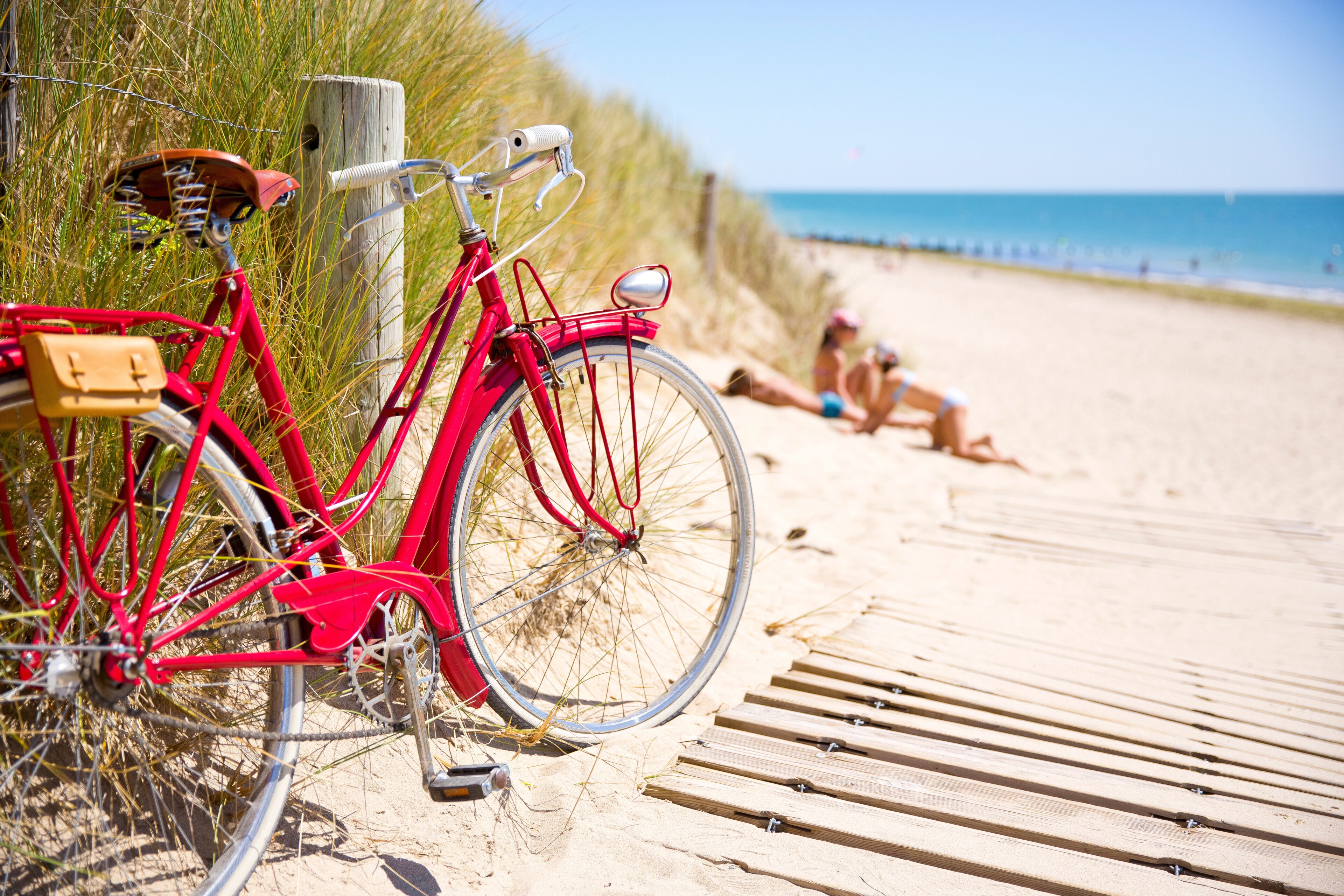 Uma bicicleta vermelha estacionada numa praia de areia, com um calçadão de madeira e um poste de madeira ao fundo.