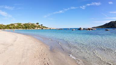 A clear beach with calm waters, rocky outcrops, and a forested hill in the background.