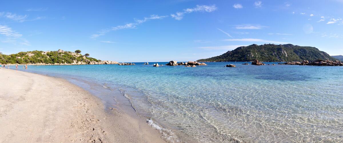 A clear beach with calm waters, rocky outcrops, and a forested hill in the background.