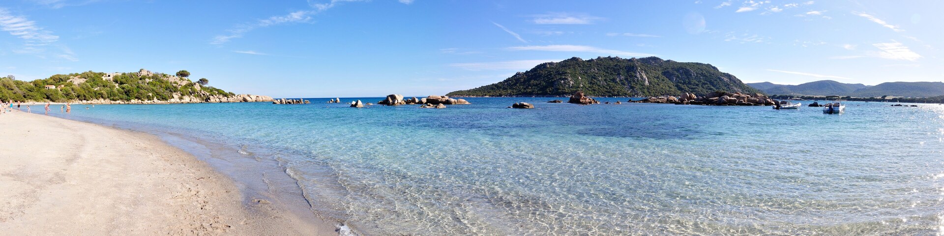 A clear beach with calm waters, rocky outcrops, and a forested hill in the background.