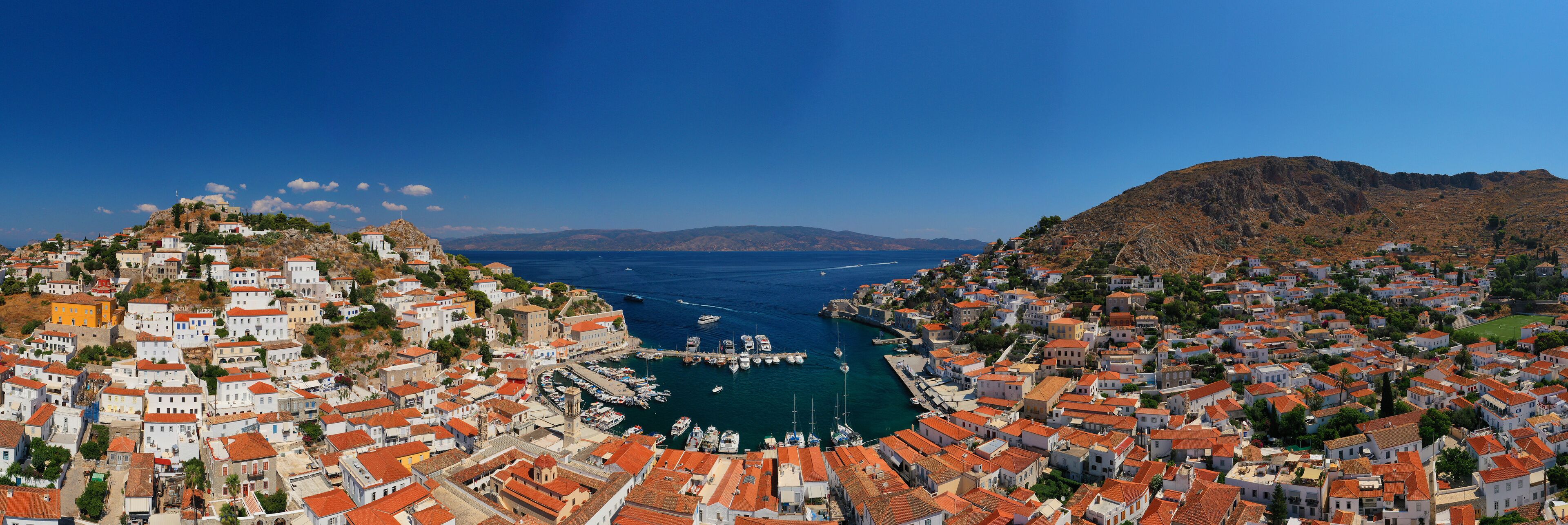 A coastal town with terracotta-roofed buildings, a marina, and a mountainous backdrop.