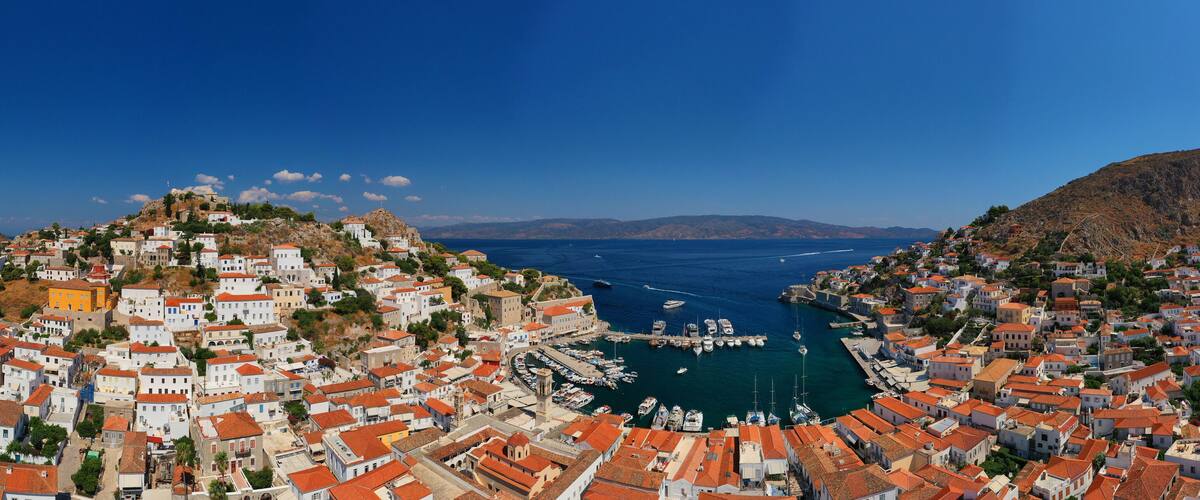 A coastal town with terracotta-roofed buildings, a marina, and a mountainous backdrop.
