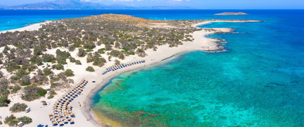 A coastal landscape with a sandy beach, clear turquoise waters, and a mountainous backdrop.
