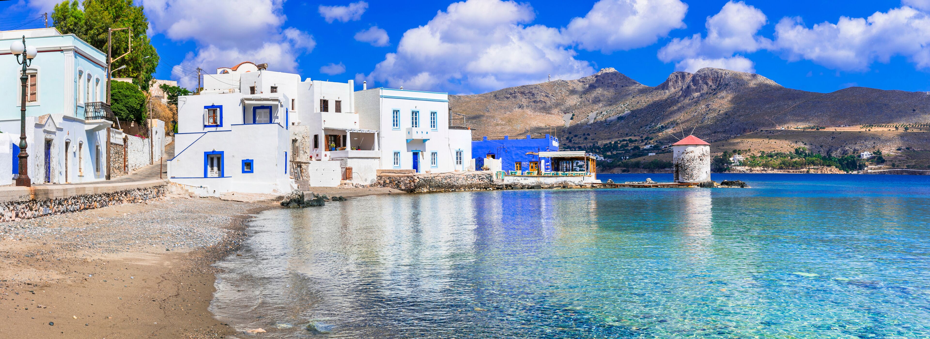 A coastal village with white buildings, a clear blue sea, and mountains in the background.