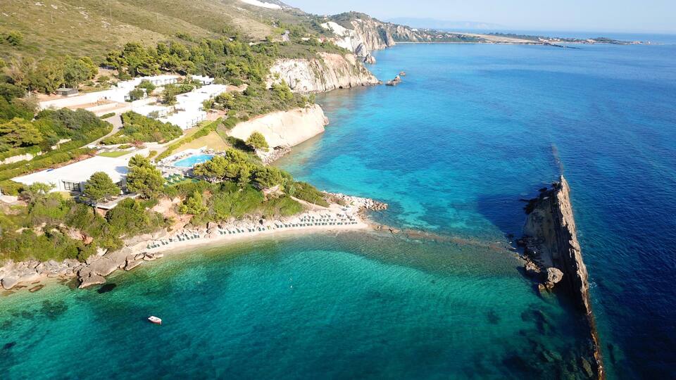 A coastal area with a clear blue sea, a rocky shoreline, and a resort with a swimming pool.