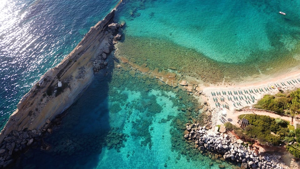A coastal landscape with clear turquoise waters, rocky cliffs, and a sandy beach with umbrellas.