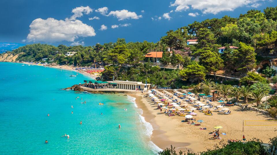 A beach with clear turquoise waters, a sandy shore, and a backdrop of lush green trees and buildings.