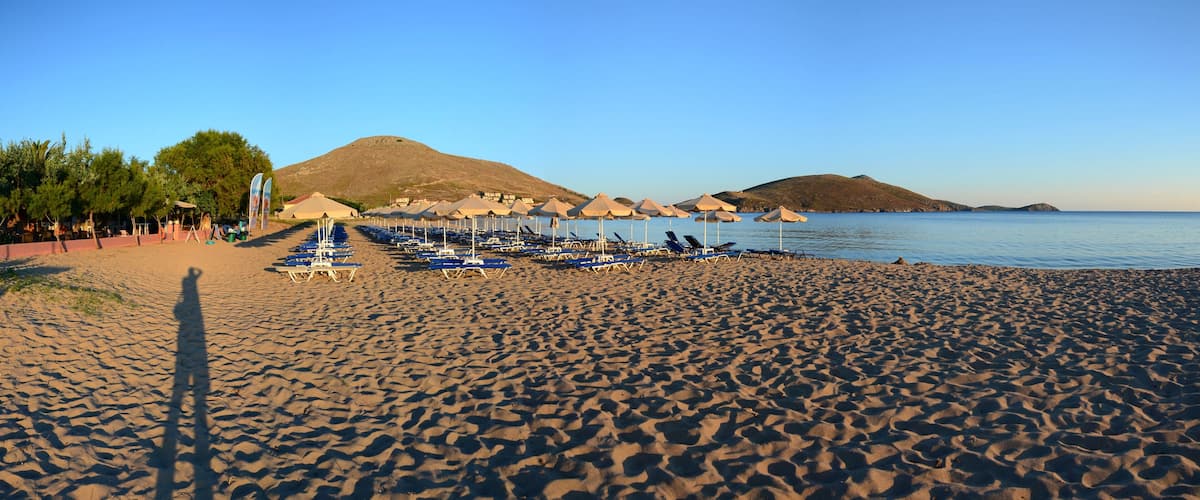 A beach with rows of sun loungers and umbrellas, a mountain in the background, and calm water.