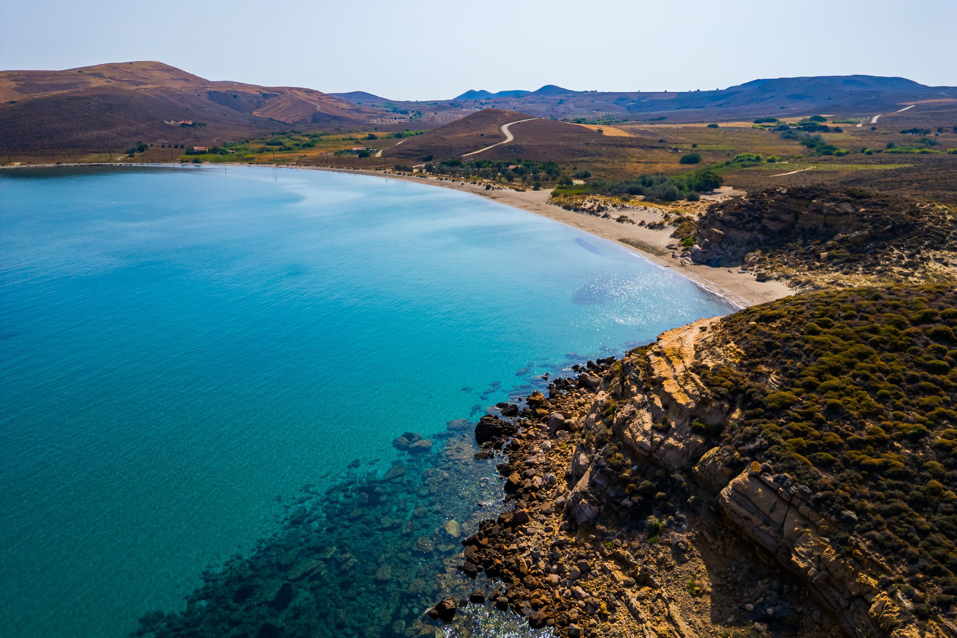 High-angle aerial view of the Neftina Beach on Limnos Island, Greece, showing crystal clear turquoise waters meeting a sandy shoreline backed by rugged, arid Mediterranean hills.