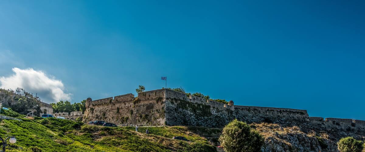 A historic stone fortress with a flag on top, surrounded by lush greenery and rocky terrain.